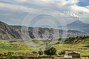 Mountain village at the foot of the Gegham ranges and the silhouette of mount Ararat, covered with haze and layered clouds in the