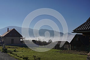 Horské vesnické domy nebo farmy v ranní mlze. dřevěné budovy se zahradou a trávníkem za slunečného dne. Odpočinková atmosféra pro turisty,