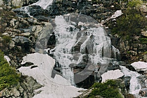 Mountain waterfall flows in turbulent stream along stone channel. Water boils between boulders, forms splashes