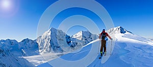 Mountaineer backcountry ski walking up along a snowy ridge with skis in the backpack. In background blue sky and shiny sun and