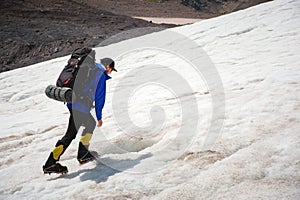 A mountaineer with a backpack walks in crampons walking along a dusty glacier with sidewalks in the hands between cracks