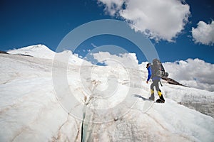 A mountaineer with a backpack walks in crampons walking along a dusty glacier with sidewalks in the hands between cracks