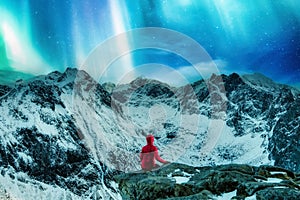 Mountaineer in red jacket sitting on the cliff with Aurora borealis over snowy mountain on Ryten in Lofoten Islands