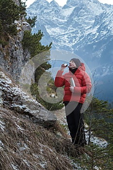 Mountaineer at winter snowy peaks background resting and drinking from vacuum flask metal cup