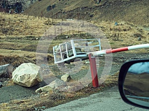 In a mountainous area among dry grass and boulders, a metal barrier gate weighted with stones blocks the road