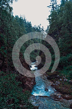 A mountainous clean blue river flows between stones rocks and mountains covered with green trees and bushes, rainy morning