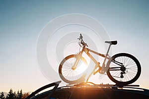 Mounted mountain bicycle silhouette on the car roof with evening sun light rays background. Safe sport items transportation using