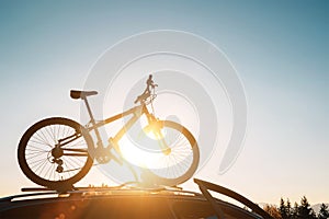 Mounted mountain bicycle silhouette on the car roof with evening sun light rays background. Safe sport items transportation using