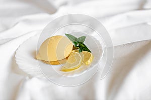 Mousse yellow cake in the form of a lemon with a green mint leaf on a white plate with a silver teaspoon on a light background.