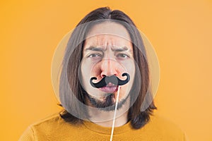 Movember concept - young frowned man holds fake paper moustache on a stick yellow background copy space isolated studio