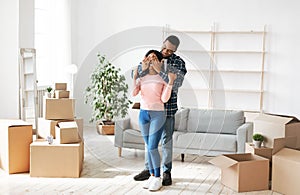 Moving day concept. Millennial black man closing his girlfriend`s eyes in their home with cardboard boxes