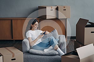 Moving day. Girl using phone app shopping furniture in online store sitting between cardboard boxes