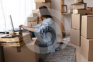 moving day, happy young woman sitting on floor and using laptop in new house or apartment among cardboard boxes, moving