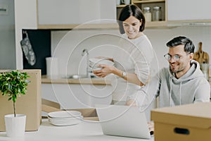 Moving house and real estate. Caring brunette woman holds pile of plates, unpacks personal belongings, husband asks her advice,