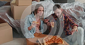 MOVING INTO NEW HOME. Young happy fun couple celebrate apartment purchase eating pizza among cardboard packaging boxes.