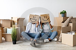 Moving and real estate concept. Black couple with cardboard boxes with smiley faces on head, sitting on floor