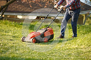 Mowing trimmer - worker cutting grass in green yard at sunset. Man with electric lawnmower, lawn mowing. Gardener