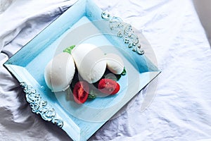 Mozzarella Italian cheese in balls with tomato and big basil leaf on square blue dish on white cotton cloth background