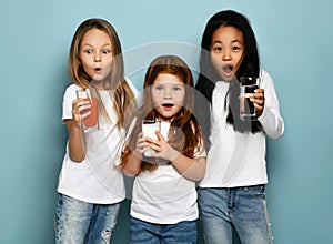 Much surprised kids girls friends in white t-shirts and blue jeans stand with glasses of water, milk and fresh juice