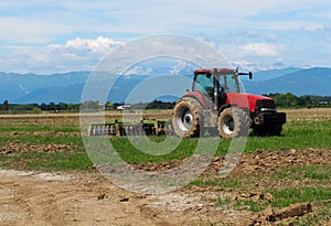 Muddy red tractor with its plow on trailer in a just plowed agricultural field. Faraway mountains on background