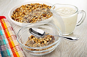 Muesli with cereals, raisin and sunflower seeds in plate, napkin, jug of yogurt, spoon and granola in bowl on wooden table