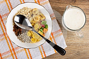 Muesli dried fruits, corn flakes, oatmeal, sunflower seeds, banana chips, spoon in bowl on napkin, jug of yogurt with cereals on