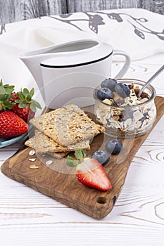 Muesli with nuts, fresh strawberries and blueberries, milk jug, crispbread on a white wooden table in a rustic style