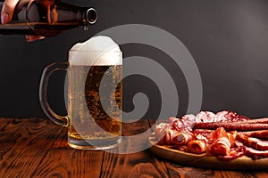 Mug of beer and assortment of snacks on a wooden table. Sausage, salami, ham, bacon. Beer salty snack, chips, wasabi nuts, salted