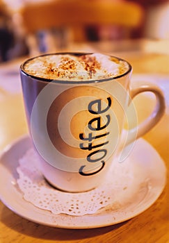 Mug of cappuccino coffee with the word coffee written on the side. On a saucer with a paper lace doily on a round wooden table