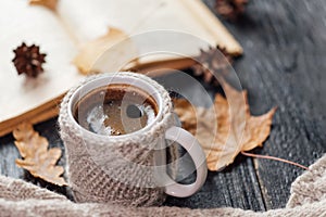 A mug of coffee in a knitted jacket, an open old book, autumn dry leaves on the table.