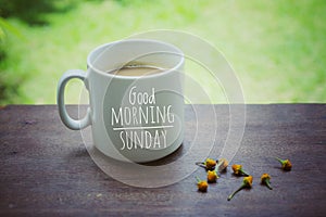 Mug of coffee. Morning white coffee with greeting text on it - Good Morning Sunday, and yellow little flowers arrangement.