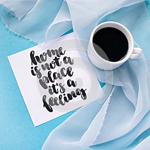 Mug of coffee on pale blue pastel chiffon cloth on a blue background. Card with handwritten inspirational quote Home is not a plac