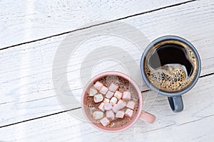 Mug of hot chocolate with small marshmallows next to a mug of black coffee isolated on white painted wood from above. Space for