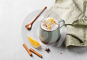 Mug of hot chocolate with whipped cream and orange zest on a light gray background with a cinnamon sticks, anise star, spoon and