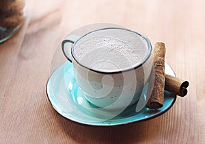 A mug of hot cocoa with froth, cinnamon sticks nearby.
