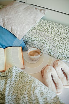 Mug of hot coffee, book, soft slippers on the bed. Breakfast in bed. Cozy home