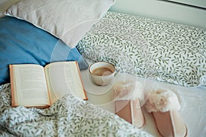 Mug of hot coffee, book, soft slippers on the bed. Breakfast in bed. Cozy home.