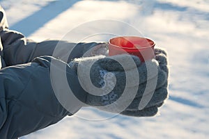 Mug with hot drink in hands dressed in knitted wool mittens in winter on a Sunny frosty day, winter picnic