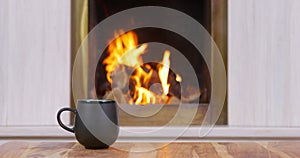 Mug with hot drink stands on table against burning fireplace