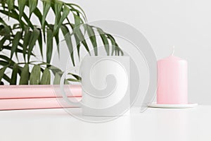 Mug mockup with books and a candle on a white table and a palm plant