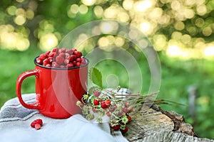 Mug and tasty wild strawberries on stump against blurred background. Space for text