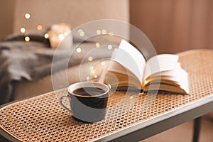 Mug of tea with open paper book on rattan coffee table in bedroom indoors close up over scented candle.