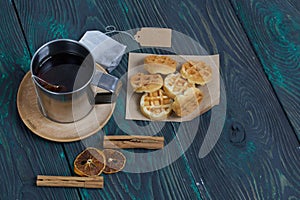 Mug of tea and waffles. Nearby is a bag of tea and spices. On painted boards. Autumn still life