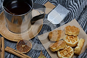 Mug of tea and waffles. Nearby is a bag of tea and spices. On painted boards. Autumn still life