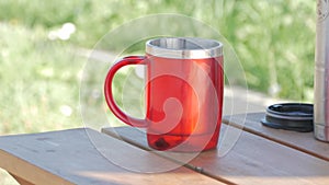 Mug thermos standing in a on a wooden table on a hot summer day. Dark red in color with black handle.