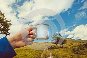 Mug of water or hot tea in hand of a lonely tourist. Hike concept in green grass mountains with dirt road.