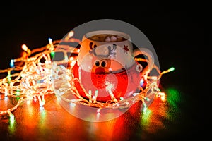 The mug is wrapped in a wool sock. A mug of cocoa and marshmallows, a garland lit next to it. Lights in the dark