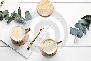 Mugs of coffee with stylish cup coasters and eucalyptus branches on white wooden table, flat lay