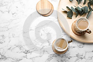 Mugs of coffee, stylish wooden cup coasters and eucalyptus branches on white marble table, flat lay. Space for text