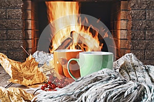 Mugs of tea or coffee, woolen thing and viburnum berries before cozy fireplace, in country house, autumn or winter holidays